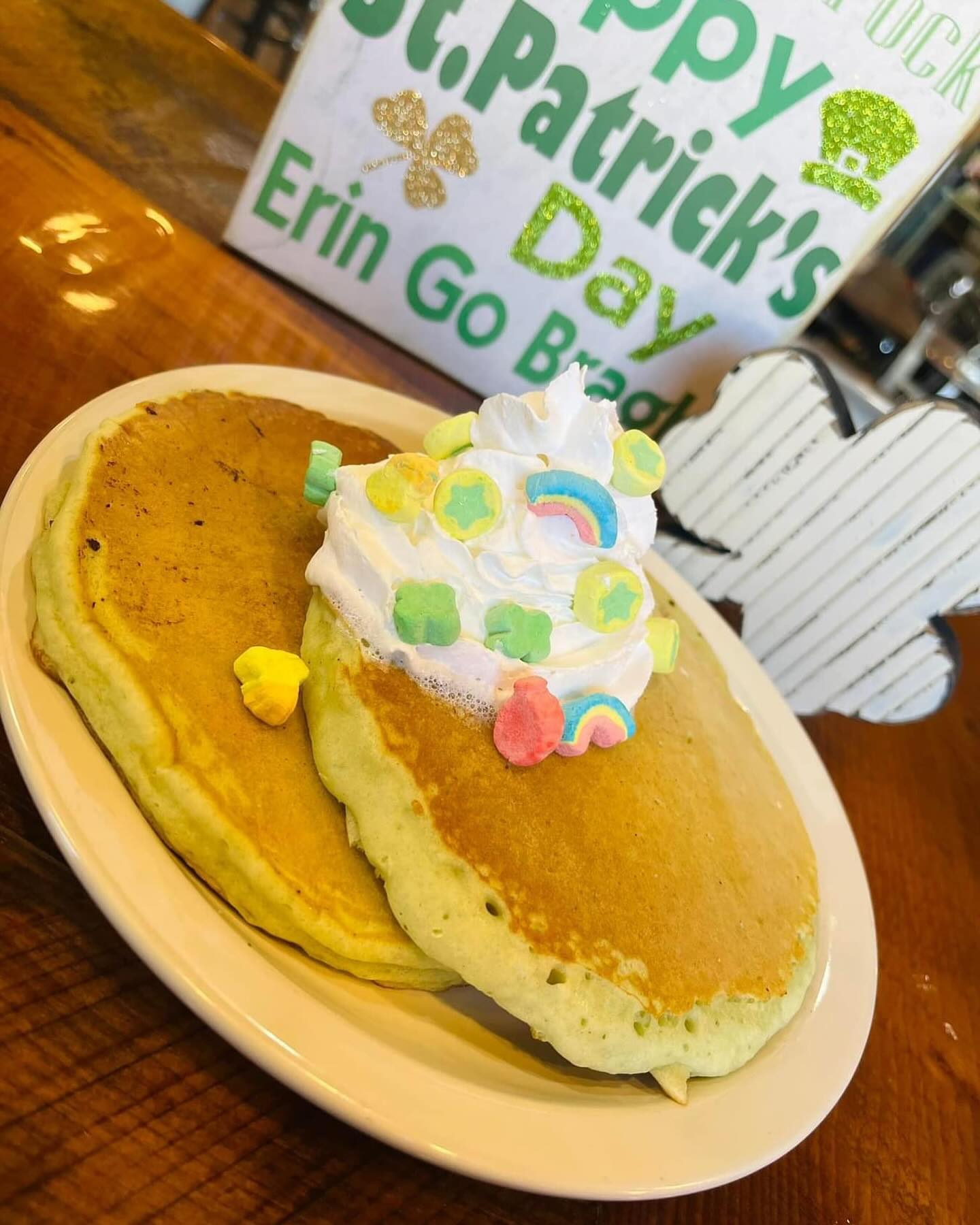 Two pancakes on a white plate topped with whipped cream and colorful St. Patrick's Day-themed candy decorations, with festive signs in the background.