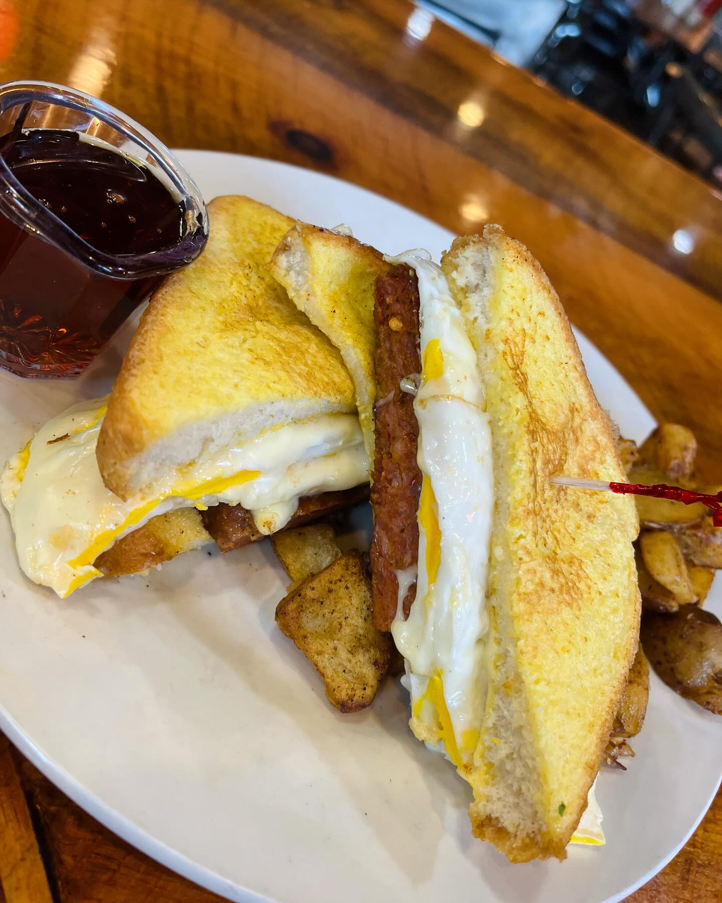 Breakfast sandwich with fried egg and sausage patties on toasted bread, served with seasoned potato wedges and a small cup of syrup on a white plate.