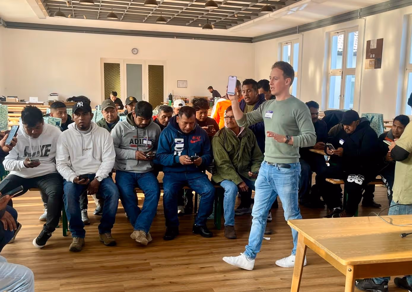 Ein Mann erklärt einer Gruppe von Menschen, die auf Bänken sitzen und auf ihre Smartphones schauen, etwas in einem hellen Raum.