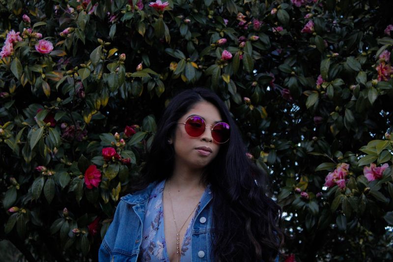 Woman with sunglasses poses in front of blooming flowers wearing a denim jacket in Gresham, OR.