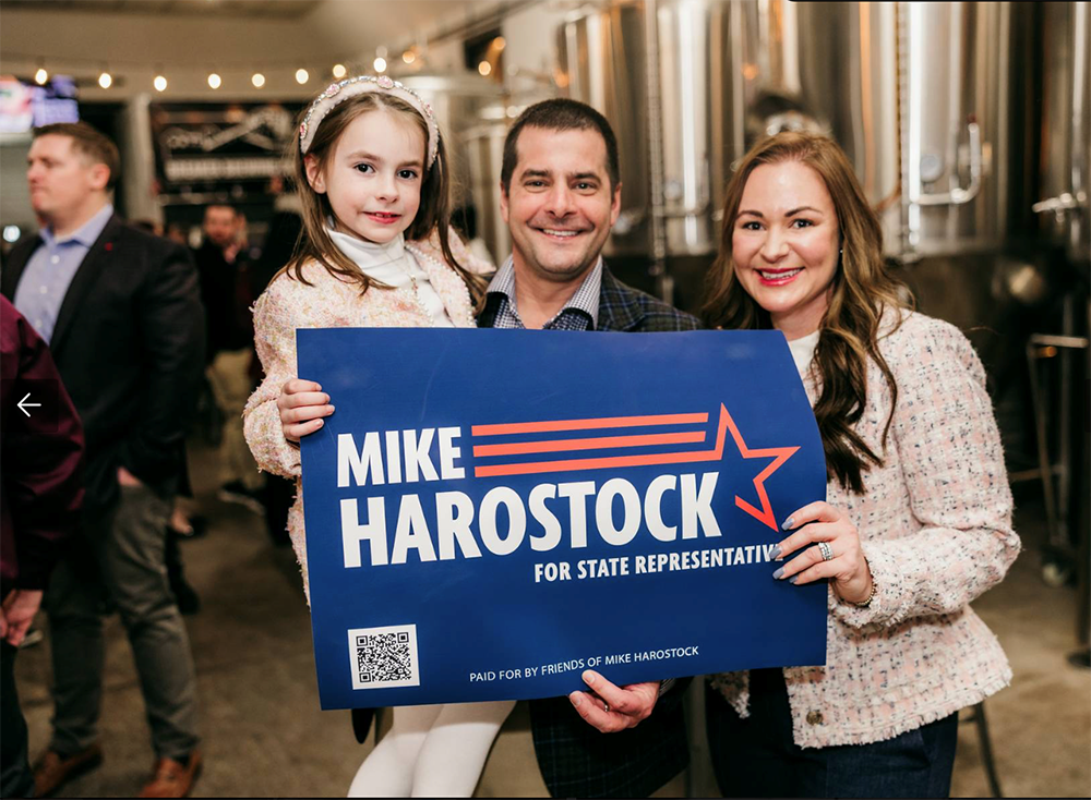 A man, woman, and young girl holding a blue campaign sign that reads 'Mike Harostock for State Representative' in an indoor setting.
