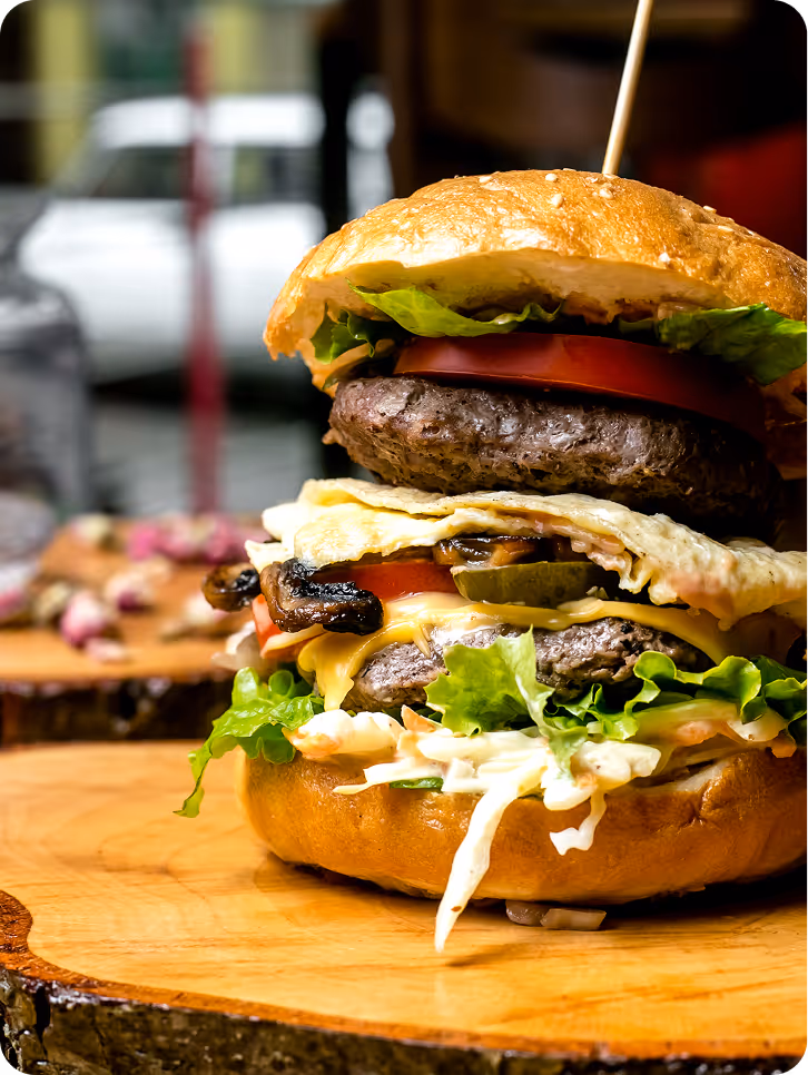 Double cheeseburger with lettuce, tomato, mushrooms, pickles, and sauce on a sesame bun, served on a wooden board.