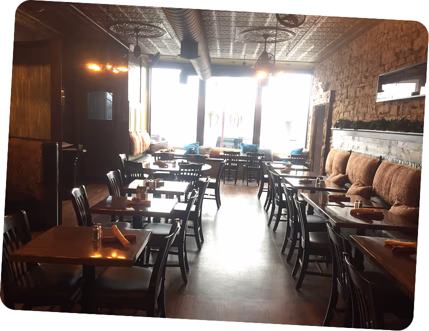 Cozy restaurant dining area with wooden tables, black chairs, cushioned bench seating along a brick wall, and large windows letting in natural light.