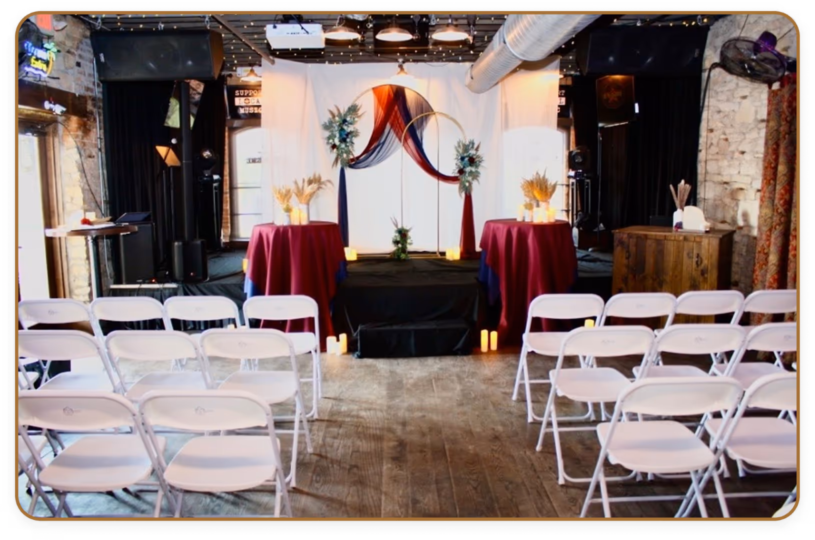 Indoor event space with white folding chairs arranged in rows facing a small stage decorated with burgundy and navy fabric, floral arrangements, and lit candles.