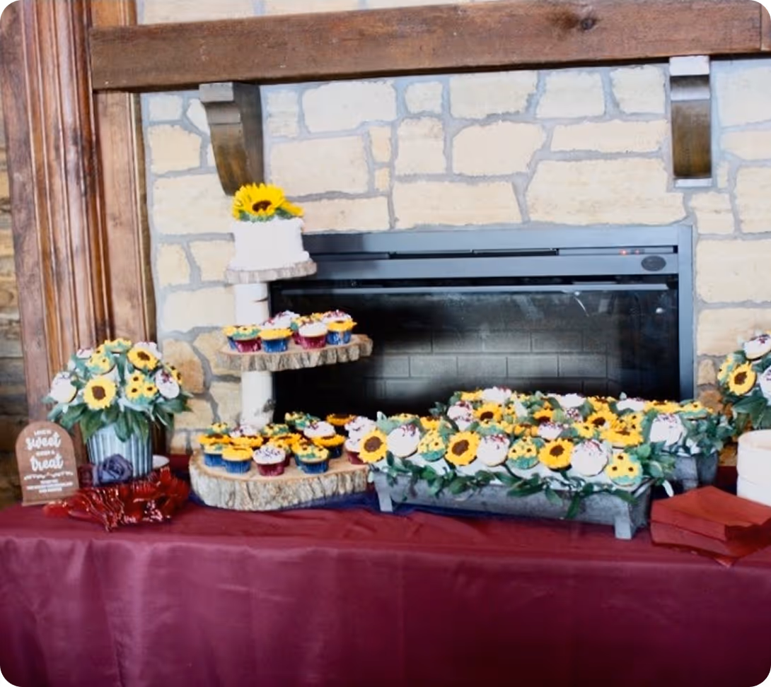 Table with sunflower-themed cupcakes and a small white cake with a sunflower on top, set against a stone fireplace backdrop.