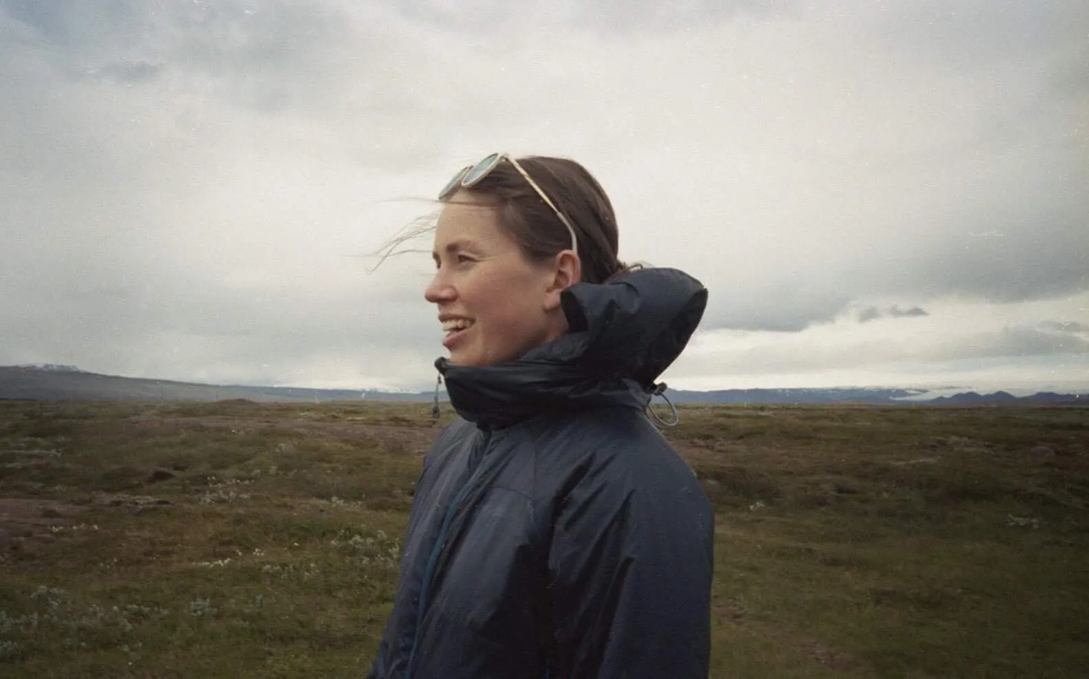 Woman in a dark jacket smiling outdoors on a cloudy day with a grassy landscape and mountains in the background.