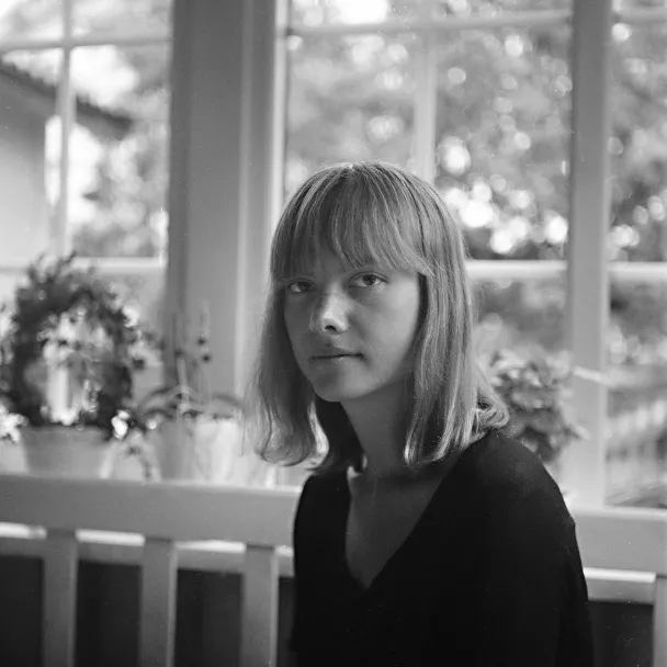 Black and white portrait of a young woman with shoulder-length hair and bangs sitting indoors near a window with potted plants.