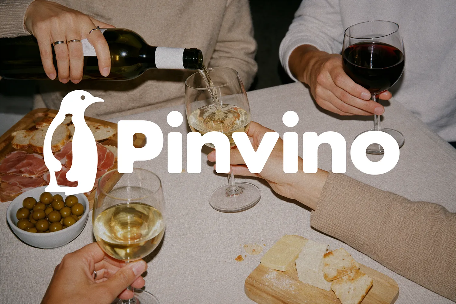 People enjoying wine with a white wine being poured into a glass and a table set with olives, cured meat, cheese, and bread.