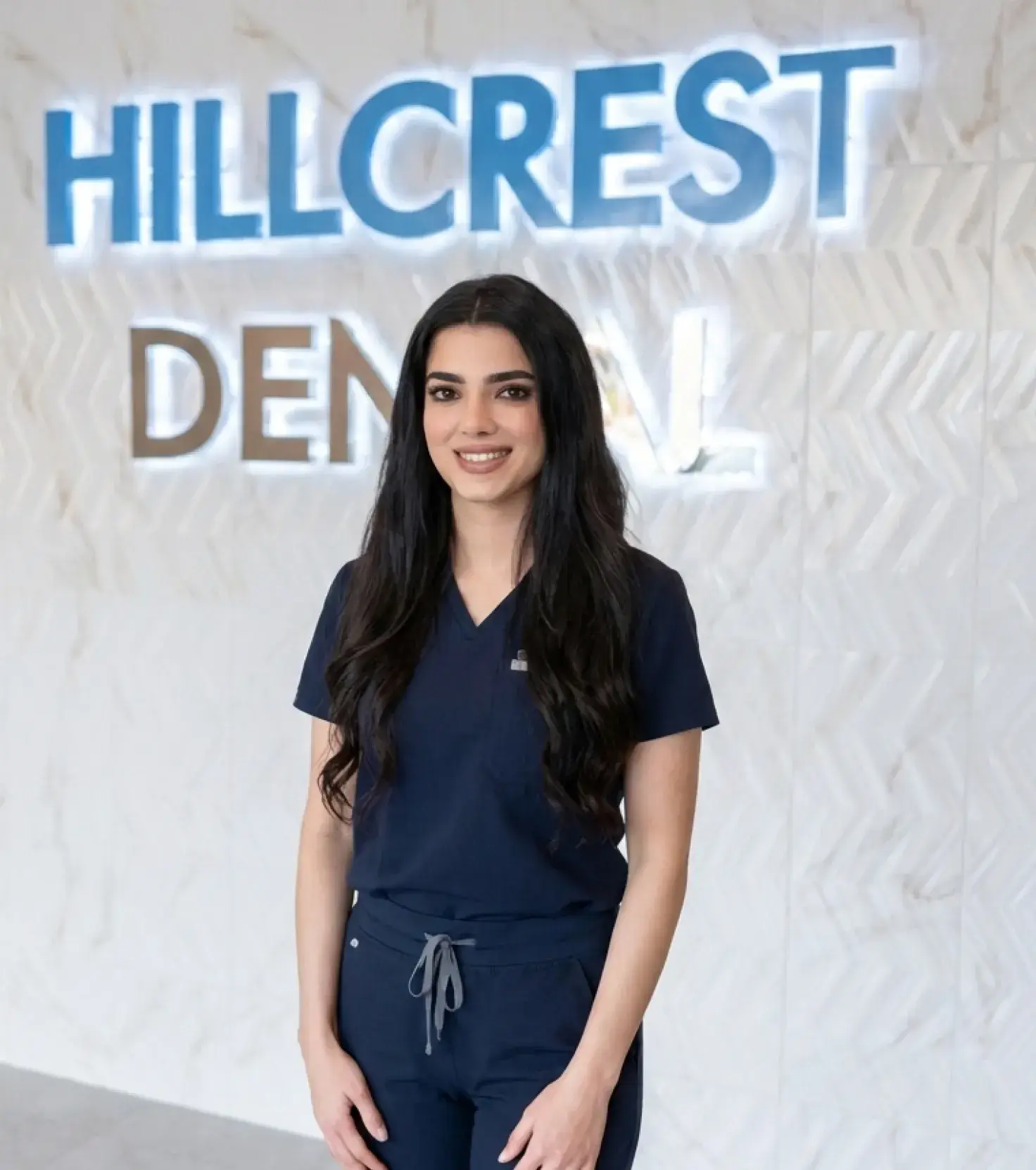 Smiling woman with long dark hair wearing navy blue medical scrubs standing in front of a Hillcrest Dental sign.
