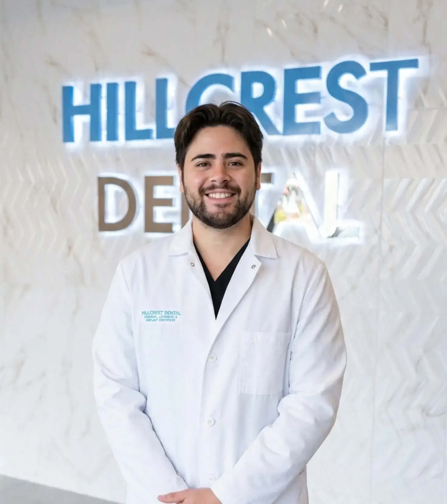 Smiling male dentist in a white coat standing in front of a wall with illuminated Hillcrest Dental sign.