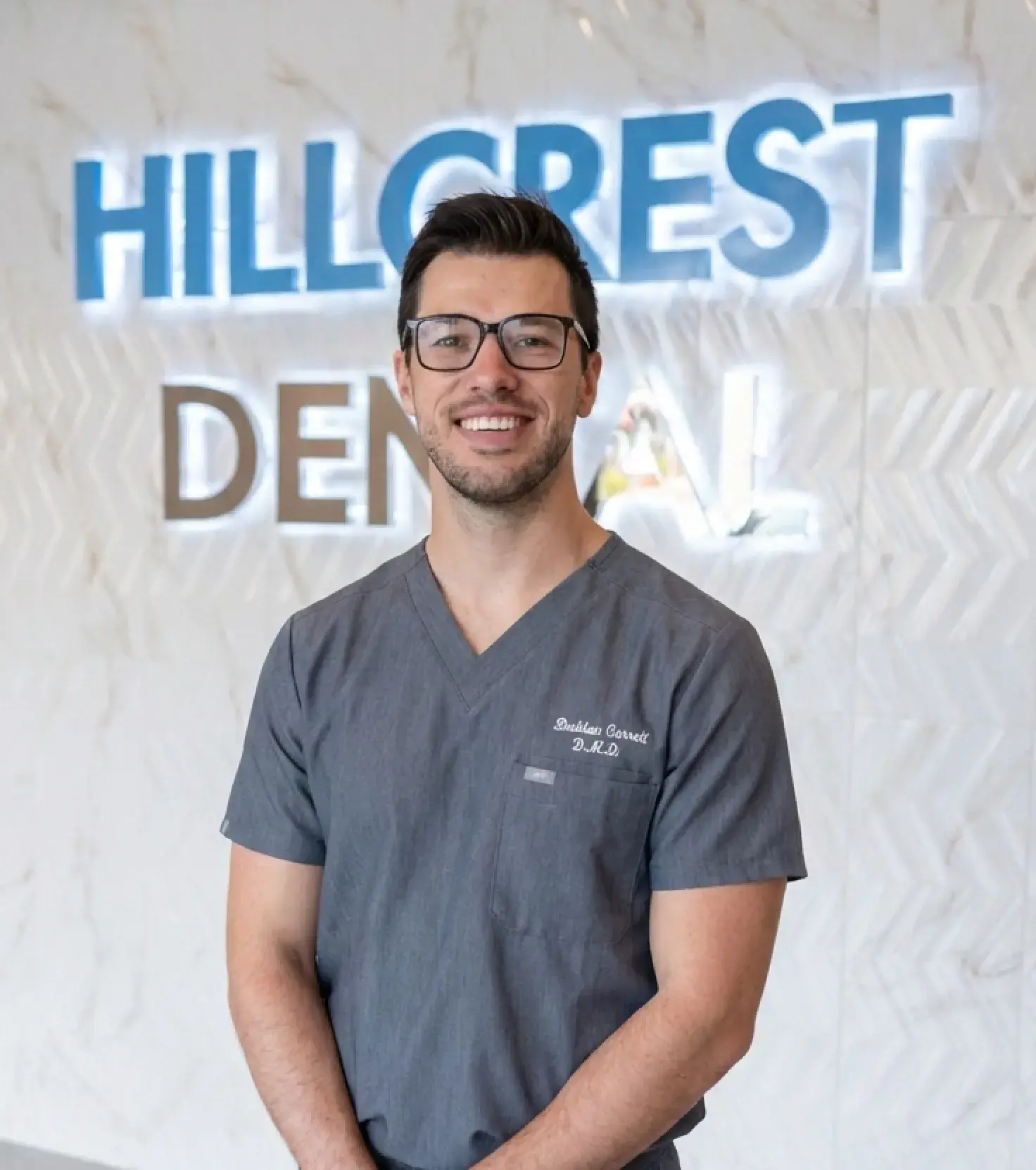 Smiling man in gray dental scrubs and glasses standing in front of a wall sign that reads Hillcrest Dental.