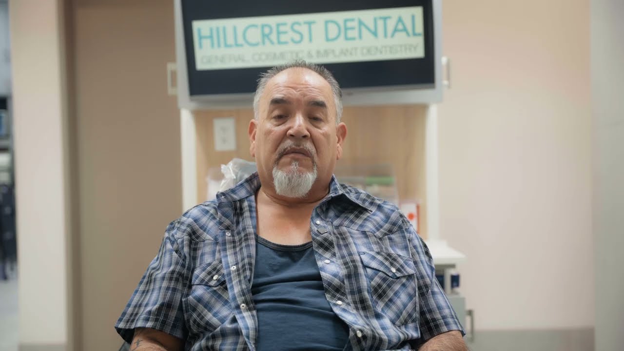 Middle-aged man with gray goatee sitting in a dental clinic chair in front of a Hillcrest Dental sign.