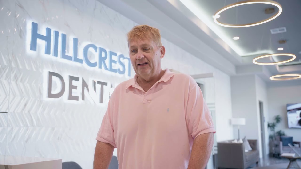 Man in a pink polo shirt standing inside a modern dental office with a lit Hillcrest Dental sign on the wall behind him.