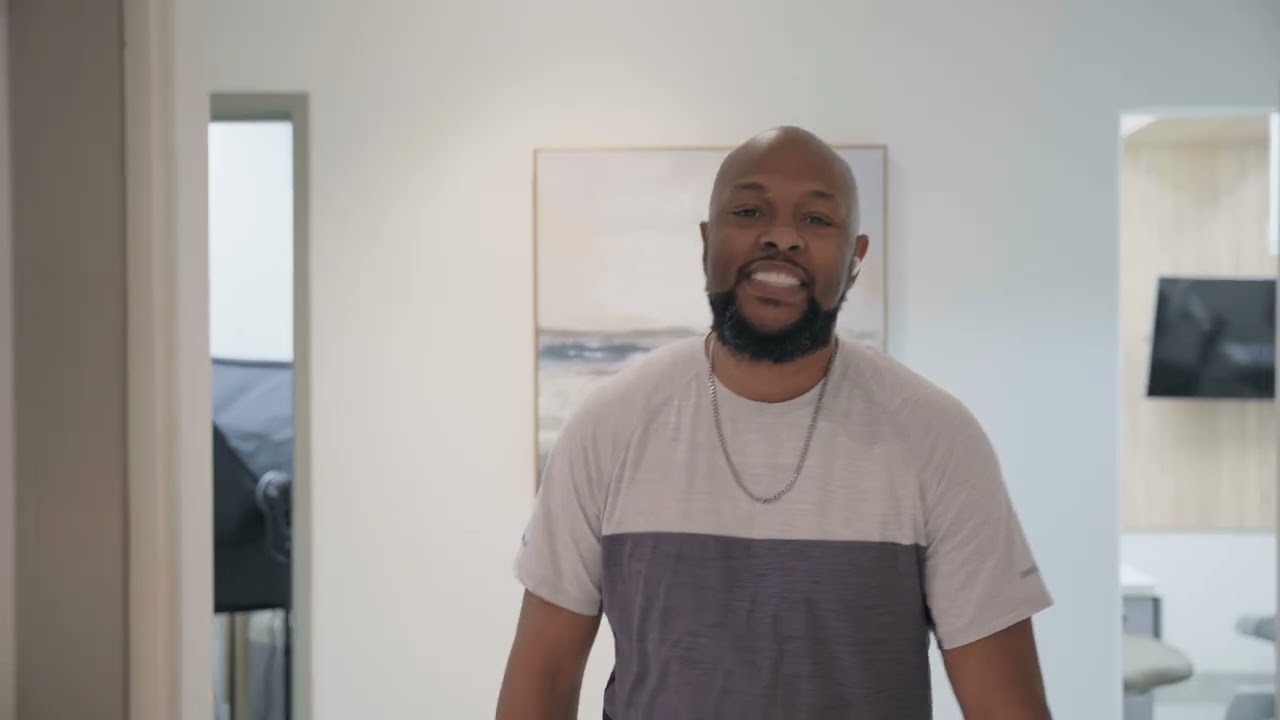 Smiling bald man with a beard wearing a two-tone t-shirt and chain necklace standing indoors.