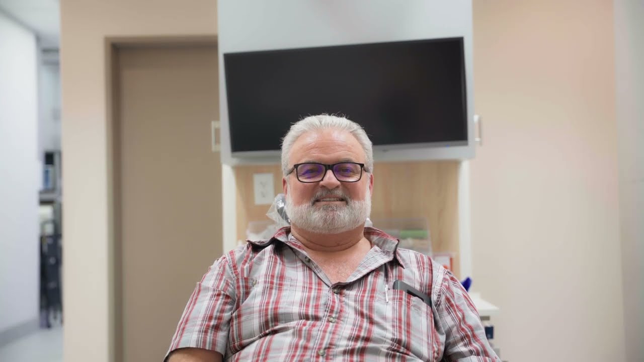 Smiling older man with white hair and beard wearing glasses and a red plaid shirt, sitting indoors in front of a blank TV screen.