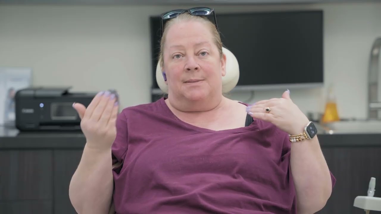 A woman with light purple nail polish wears a purple shirt and an Apple Watch, gesturing with both hands while seated in an indoor office setting.