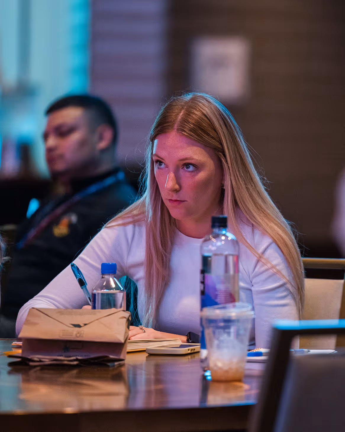 A focused young woman with long blonde hair writing notes at a table with water bottles and a paper bag, with a blurred person in the background.