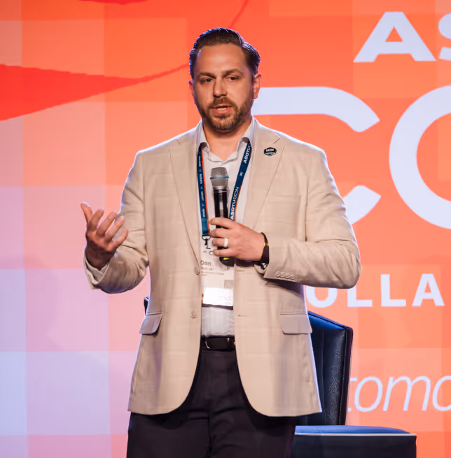 Man in beige blazer speaking into a microphone on stage with an orange background.