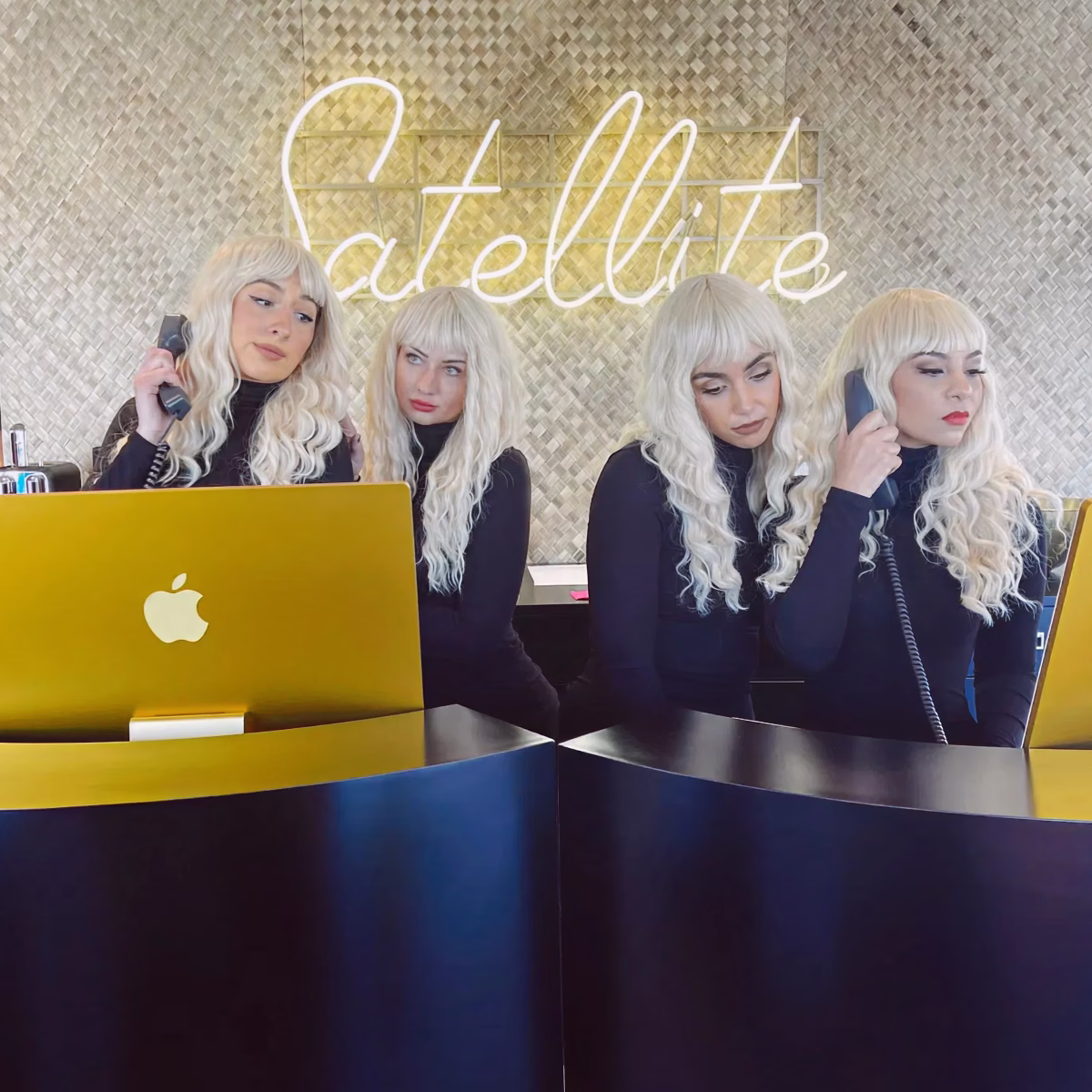 Four models in white wigs behind a yellow computer beneath the Satellite sign