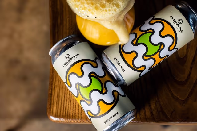 Two cans of Crooked Run Fermentation Sticky Rice beer on a wooden surface beside a glass of foamy beer.