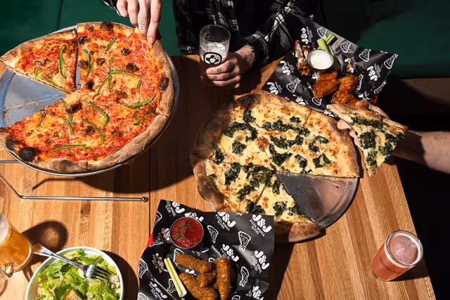 Top-down view of two pizzas, mozzarella sticks with dipping sauce, a salad, and drinks on a wooden table with two hands reaching for food.