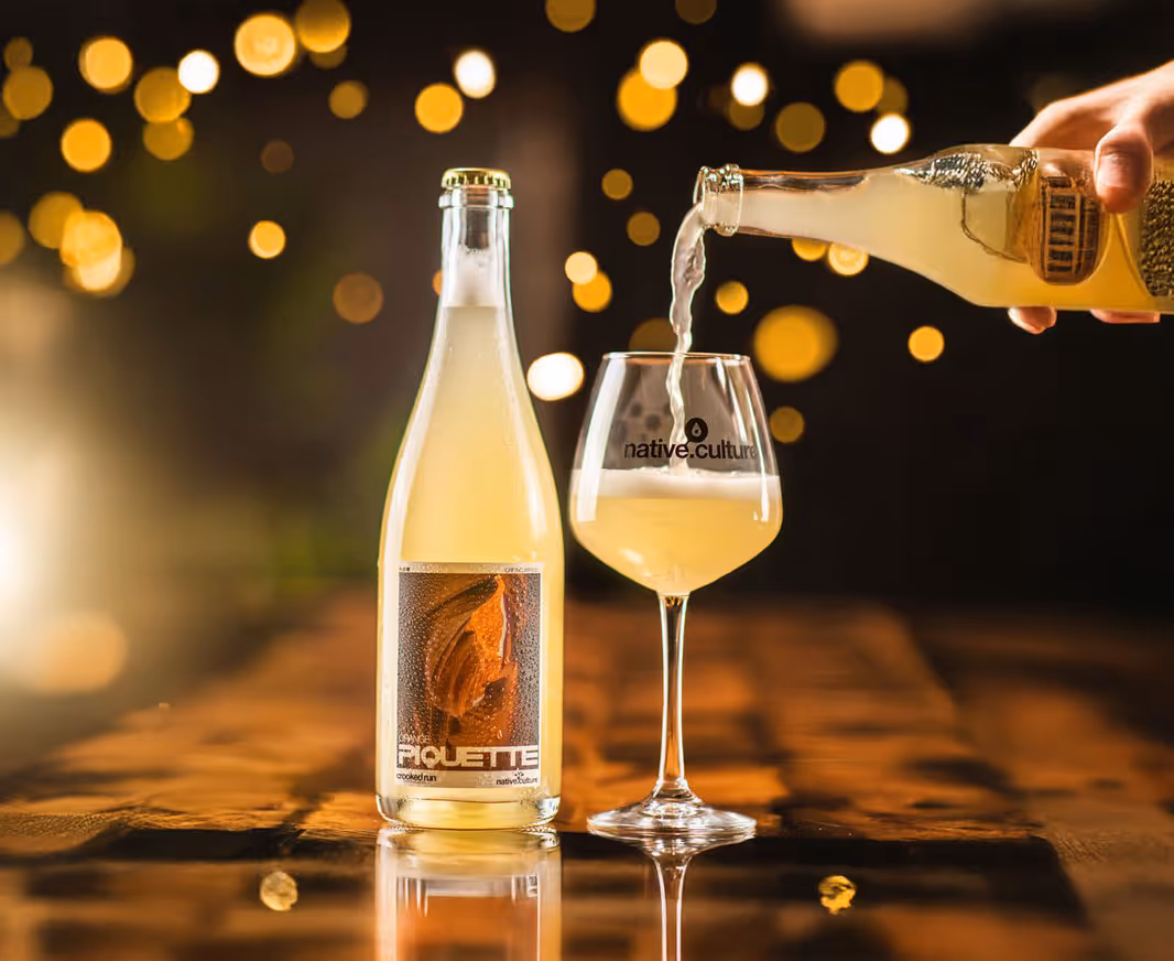 A bottle stands on a wooden table beside a glass being filled with a pale drink. Warm bokeh lights create a cozy, elegant ambiance.