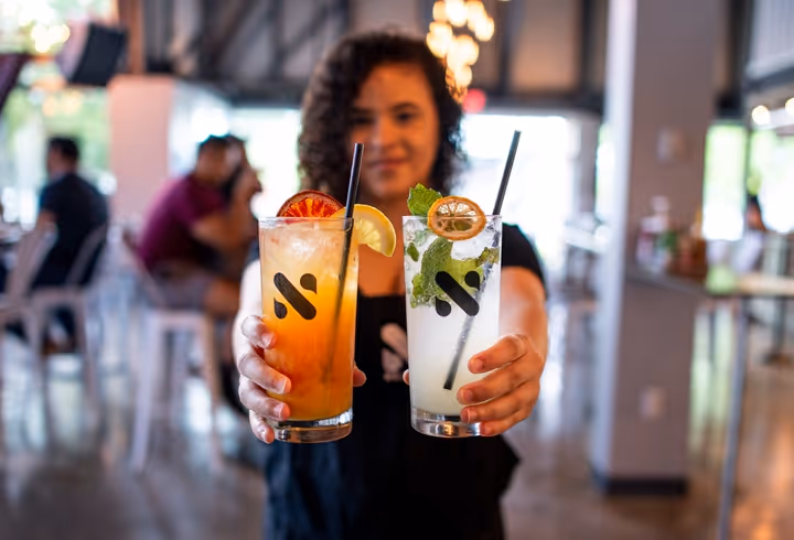 Person holding two cocktails with garnishes of lemon, blood orange, and mint in a bar setting.