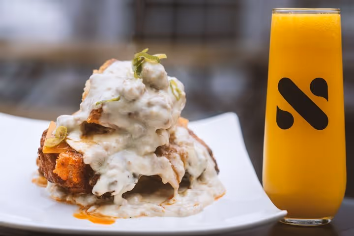Plate of biscuits and gravy with sausage and green garnish next to a glass of orange juice with a black logo.