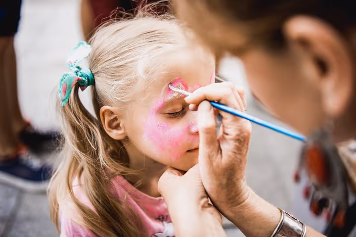Young girl with blonde hair in a ponytail having her face painted pink by an artist with a brush.