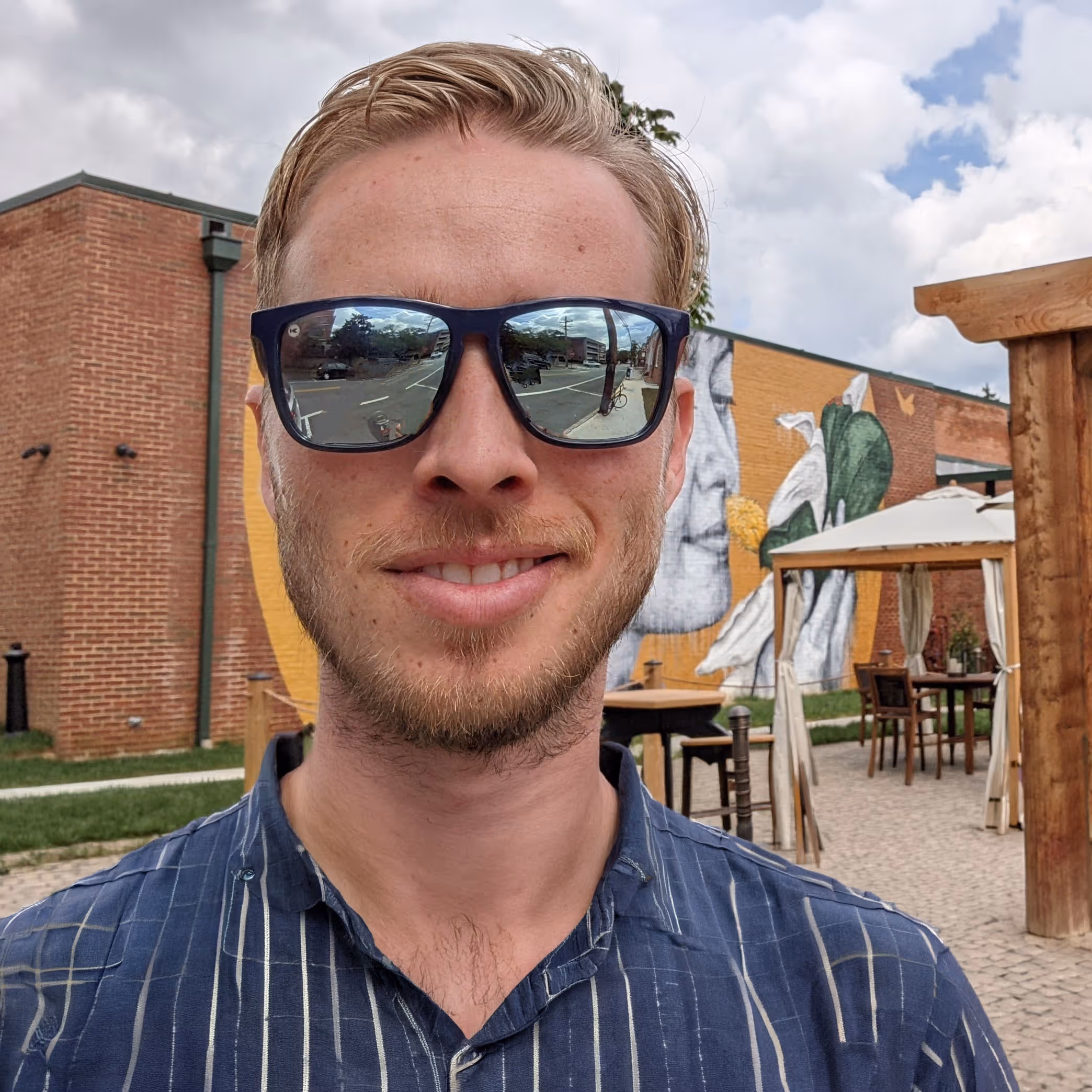 Smiling man with sunglasses reflecting a street scene, standing outdoors near a mural and patio furniture.