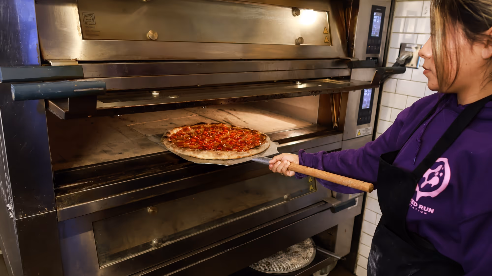 Person using a wooden pizza peel to slide a pepperoni pizza into a commercial pizza oven.