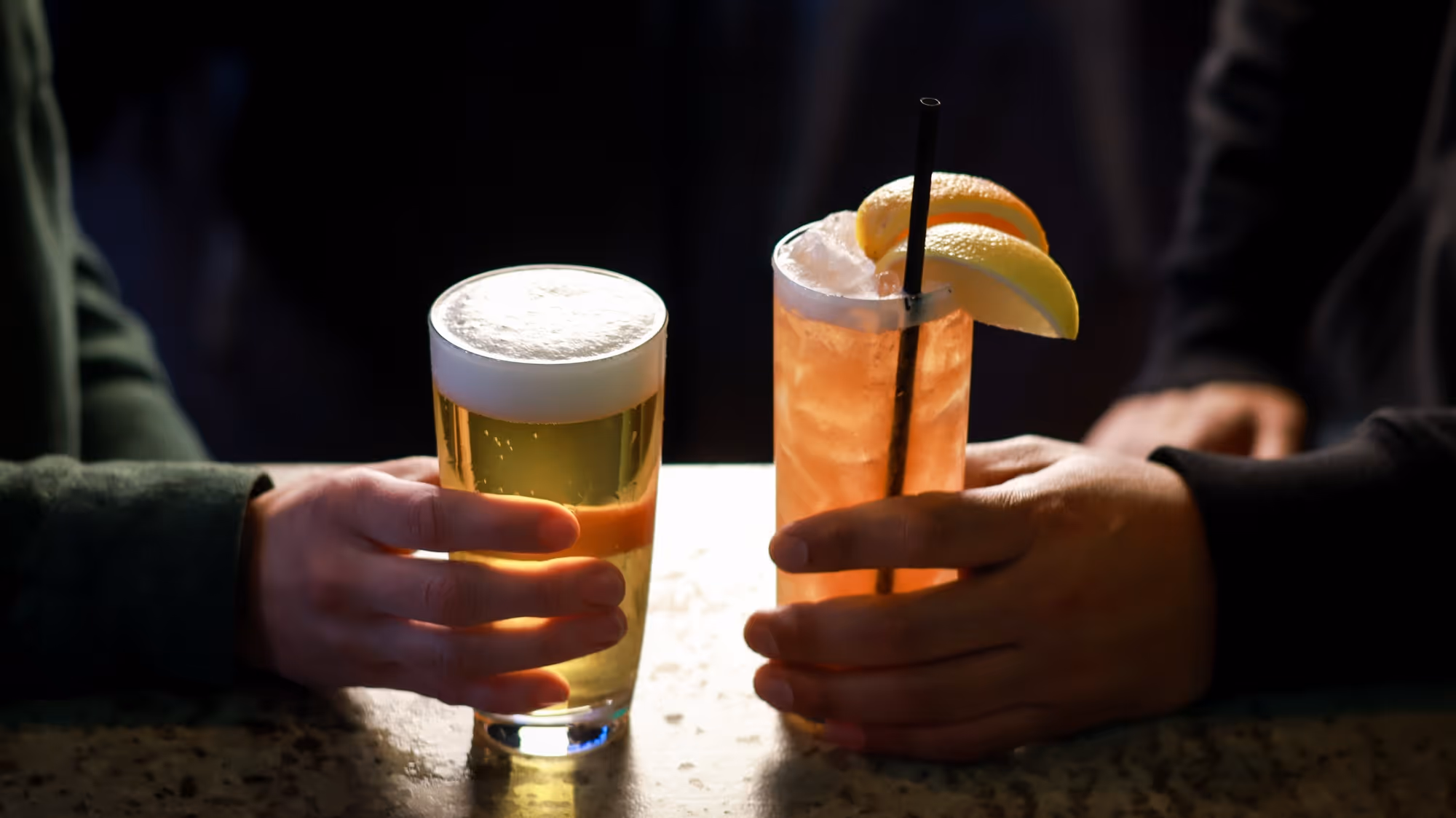Two hands each holding a different drink: one a foamy beer in a pint glass, the other an iced cocktail garnished with lemon and orange slices and a black straw.