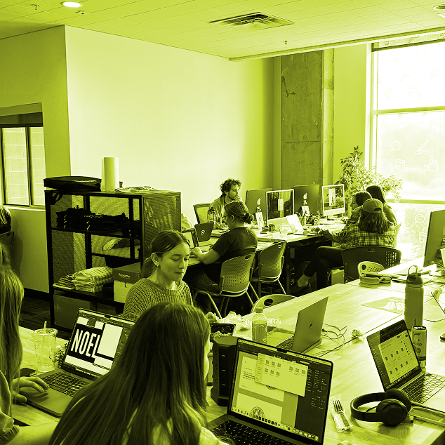 Open office space with multiple people working on laptops and desktop computers at desks near a large window.
