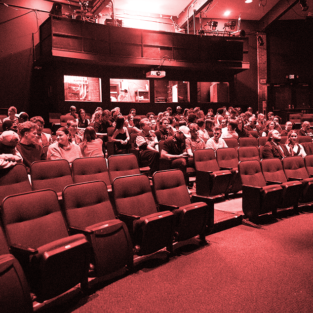 Audience seated and waiting inside a theater with red lighting and a projection booth in the background.