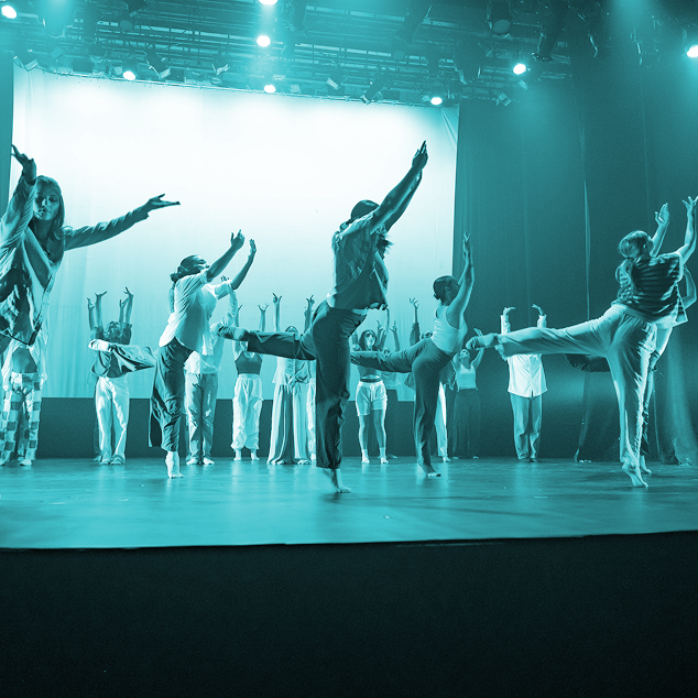Group of dancers performing on stage with arms raised and one leg lifted, illuminated by stage lights.