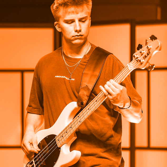 Young man wearing a shirt and necklace playing a white bass guitar indoors.