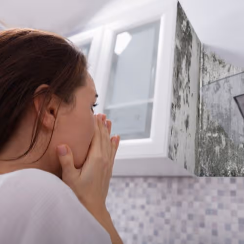 Woman covering her mouth with hands looking at a bathroom mirror with visible mold and mildew stains.