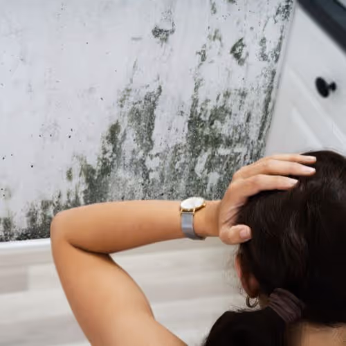 Person with dark hair holding their head and looking out a window with mold and dirt on the glass.