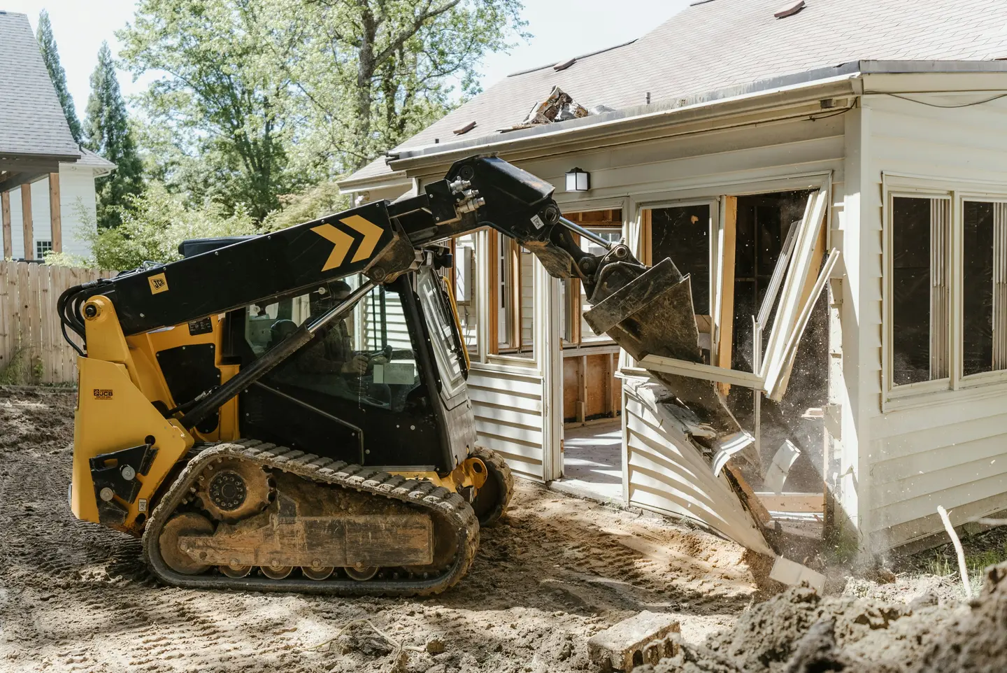 Small yellow and black excavator demolishing the siding and window frame of a white house.