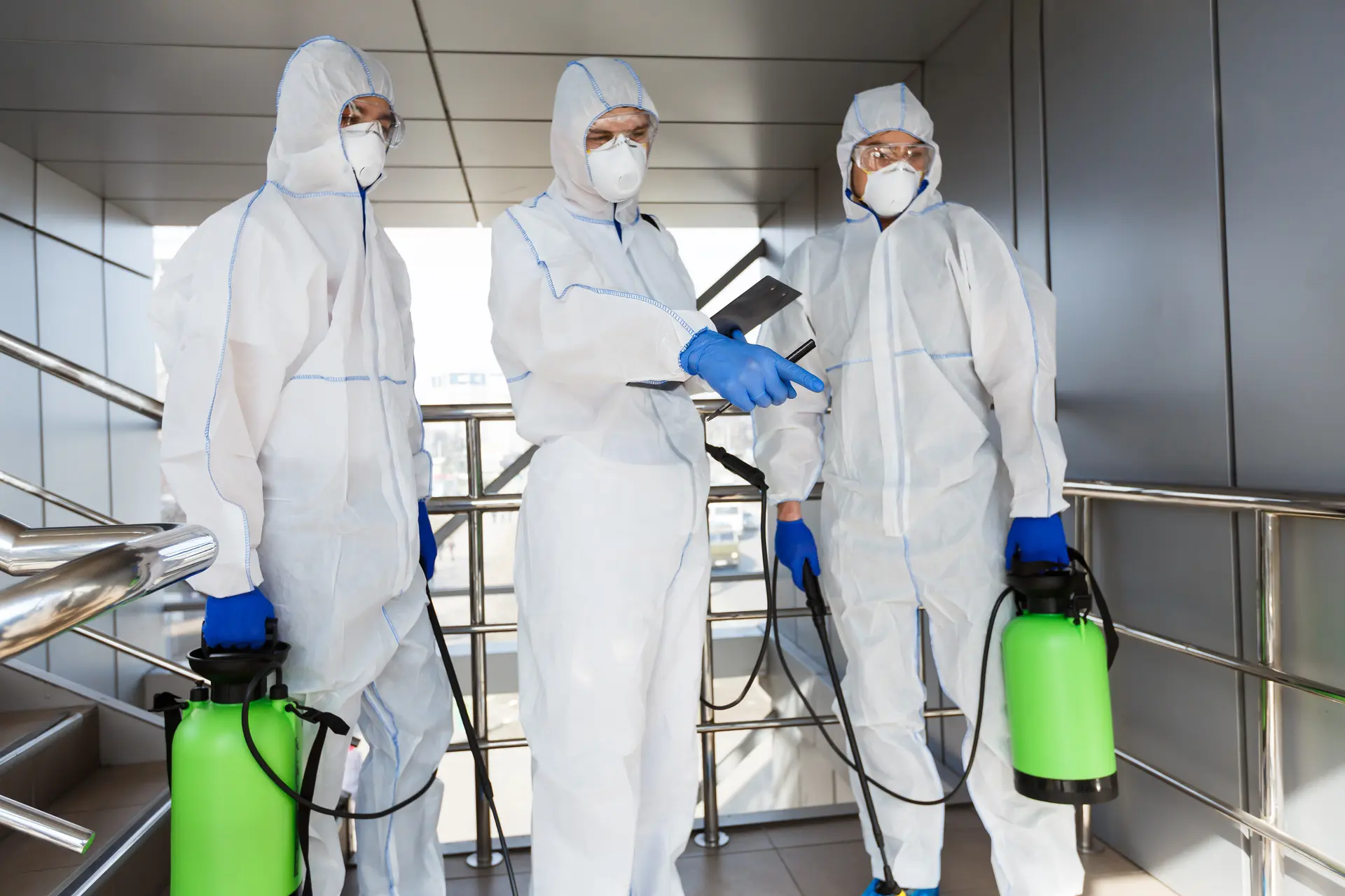 Three people in white hazmat suits, blue gloves, masks, and goggles holding green sprayers standing indoors by a stainless steel railing.