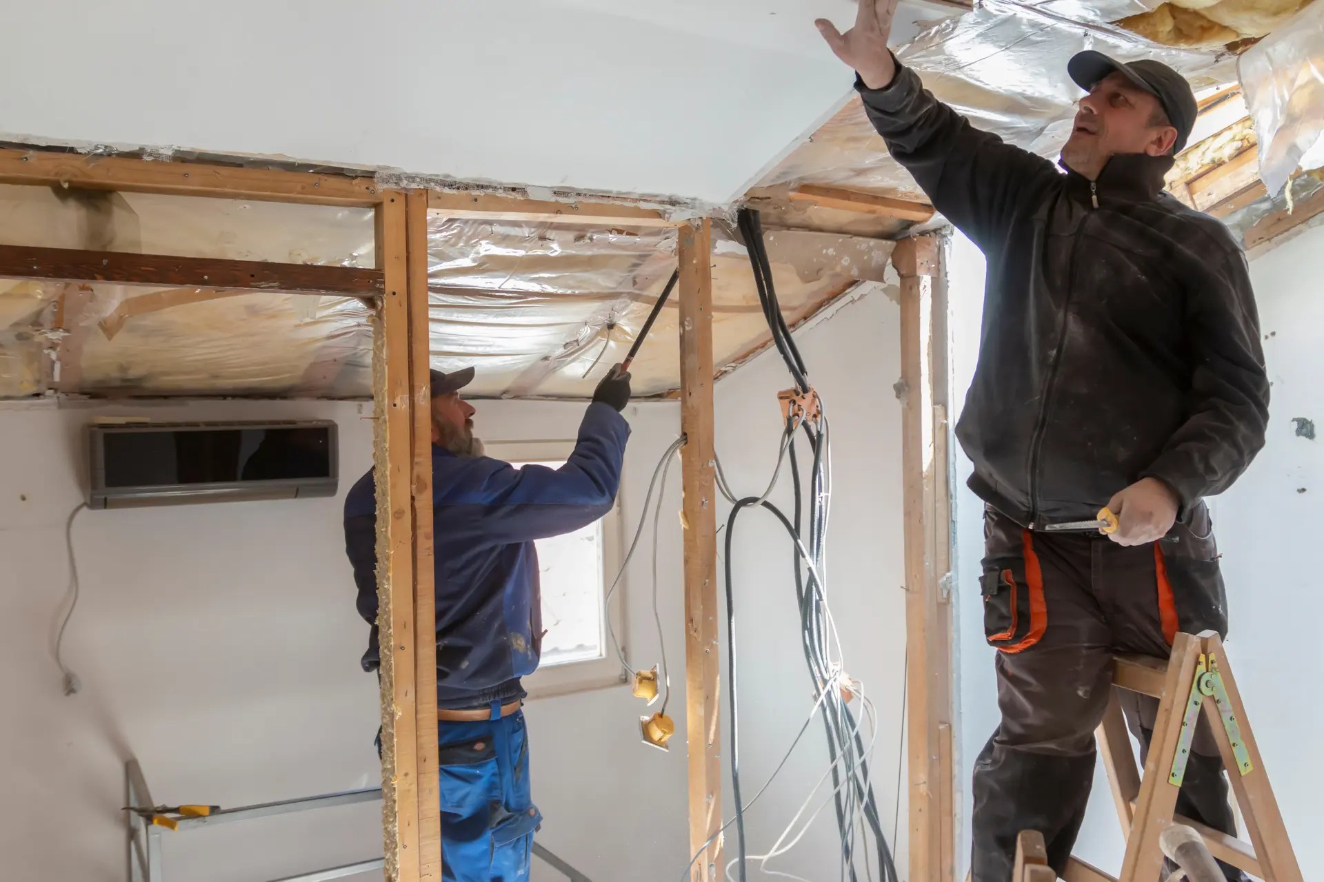 Two construction workers renovating a ceiling with exposed wiring and insulation, one on a ladder and the other using a tool.