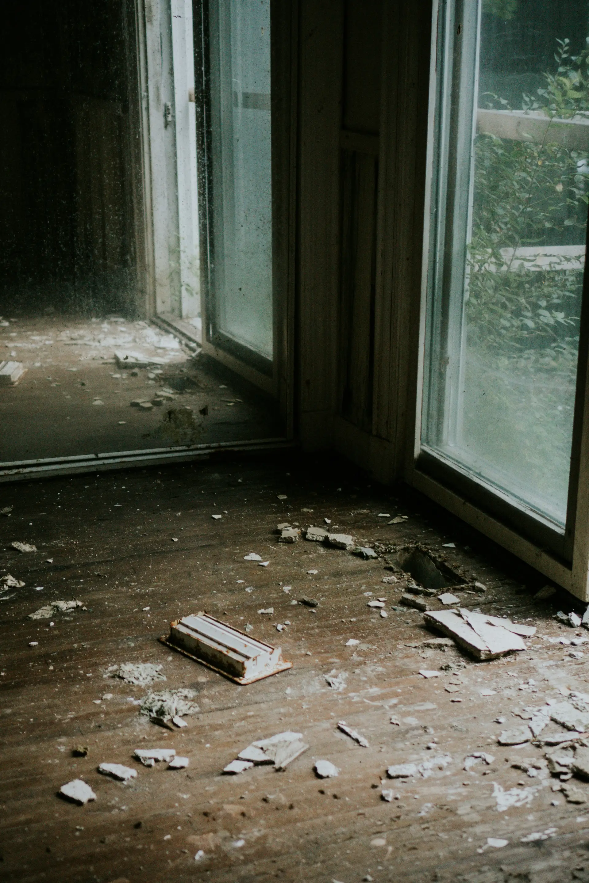 Dusty wooden floor with scattered debris and broken drywall pieces near a glass door with a reflection of the room.