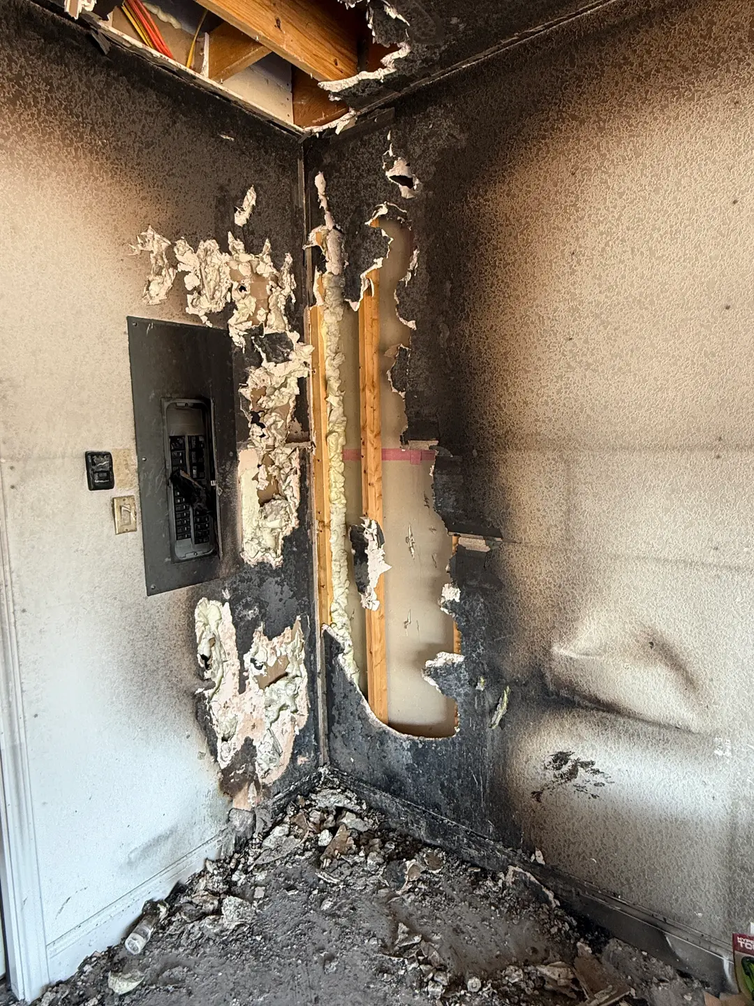 Interior corner of a room with severe fire damage, showing charred and peeling walls, exposed wooden studs, damaged electrical panel, and debris-covered floor.