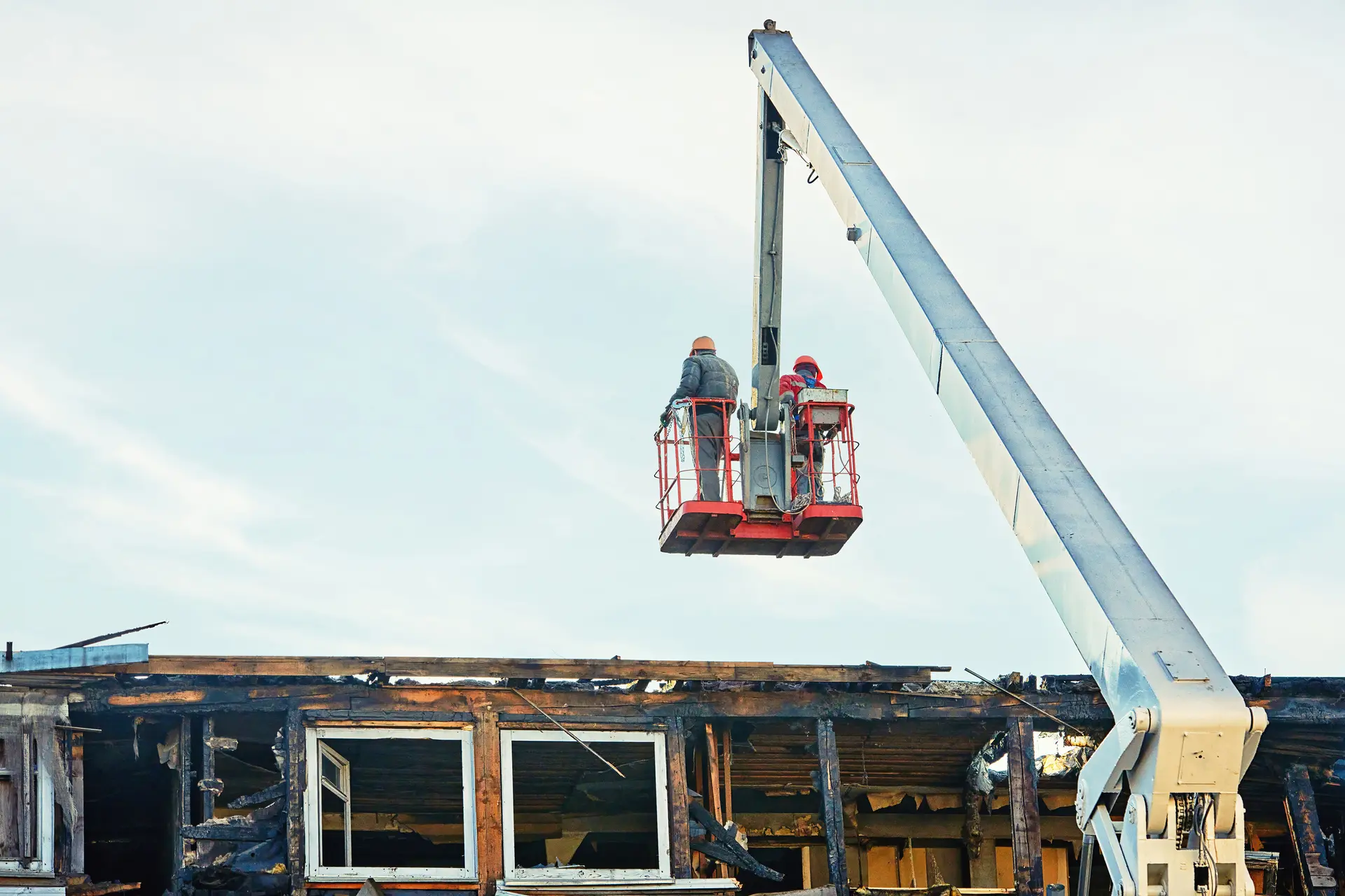 Two workers wearing helmets standing on a raised aerial lift platform inspecting a burnt, partially destroyed building.