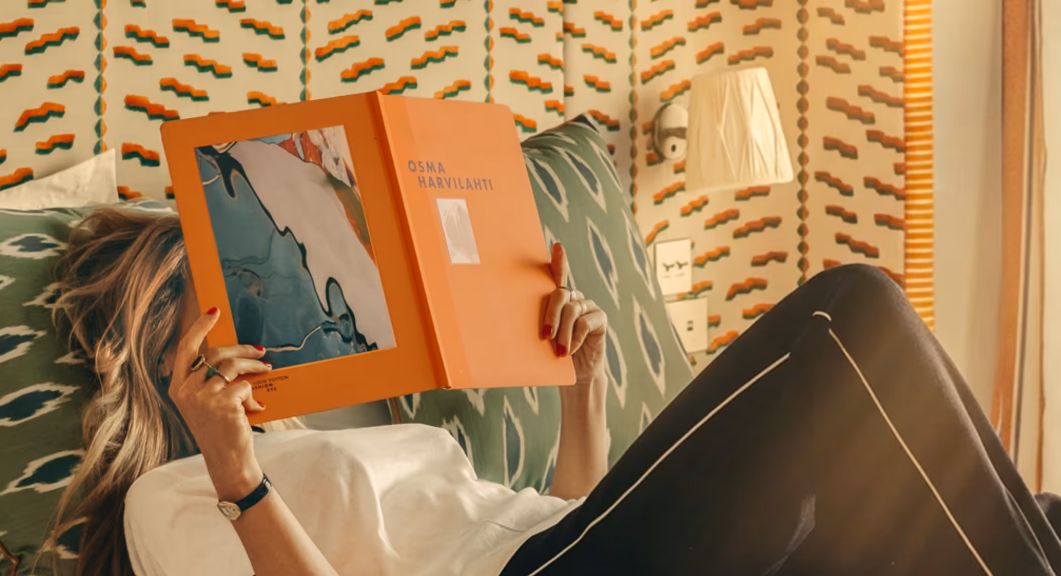 Personne allongée sur un canapé à motifs verts, lisant un livre orange nommé Osma Harvilahti.