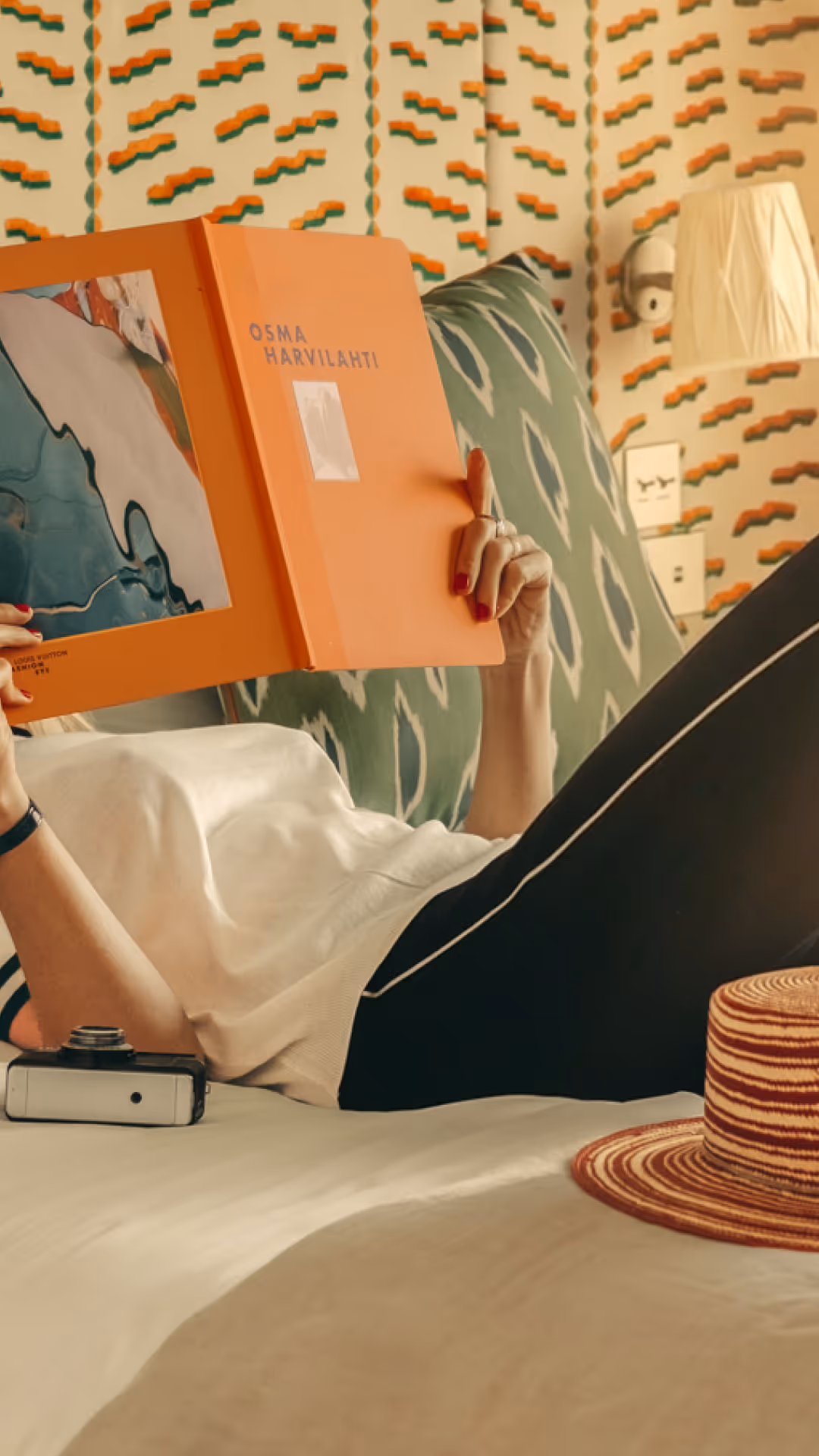 Personne allongée sur un lit lisant un livre orange intitulé Osma Harvilahti, avec un chapeau rayé posé à côté.