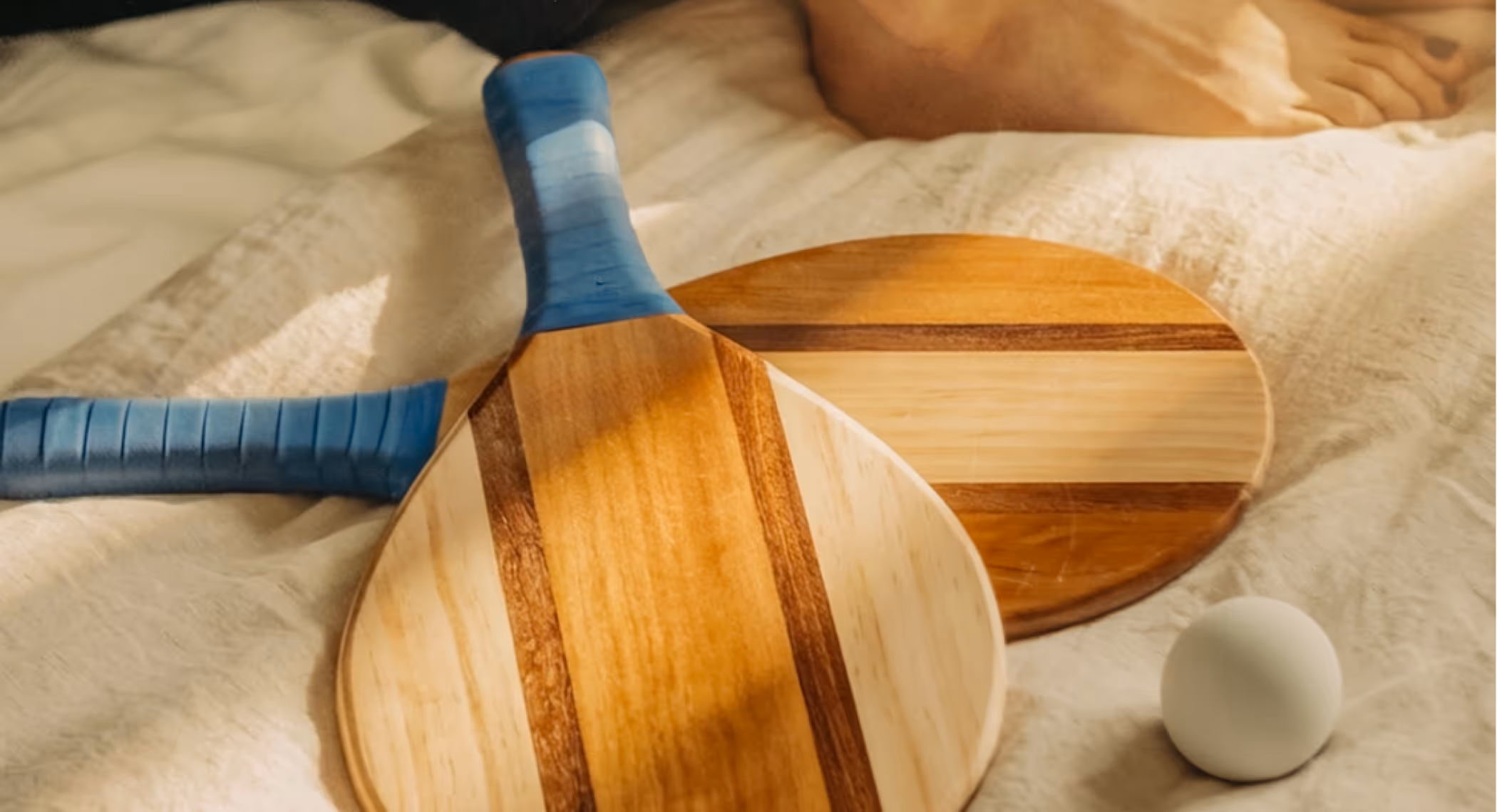 Deux raquettes de plage en bois avec poignées bleues posées sur un tissu clair, avec une balle blanche et des pieds nus visibles en arrière-plan.