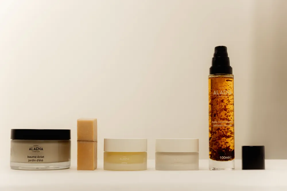 Cinq produits de soins de la marque ALAENA alignés, comprenant un baume, deux pots de crème, un sérum avec des pétales de rose visibles à l'intérieur et un bouchon noir.