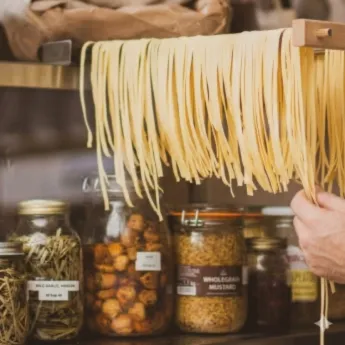 Des pâtes fraîches longues suspendues sur une étagère en bois au-dessus de bocaux en verre remplis d'herbes, champignons et condiments dans une cuisine rustique.