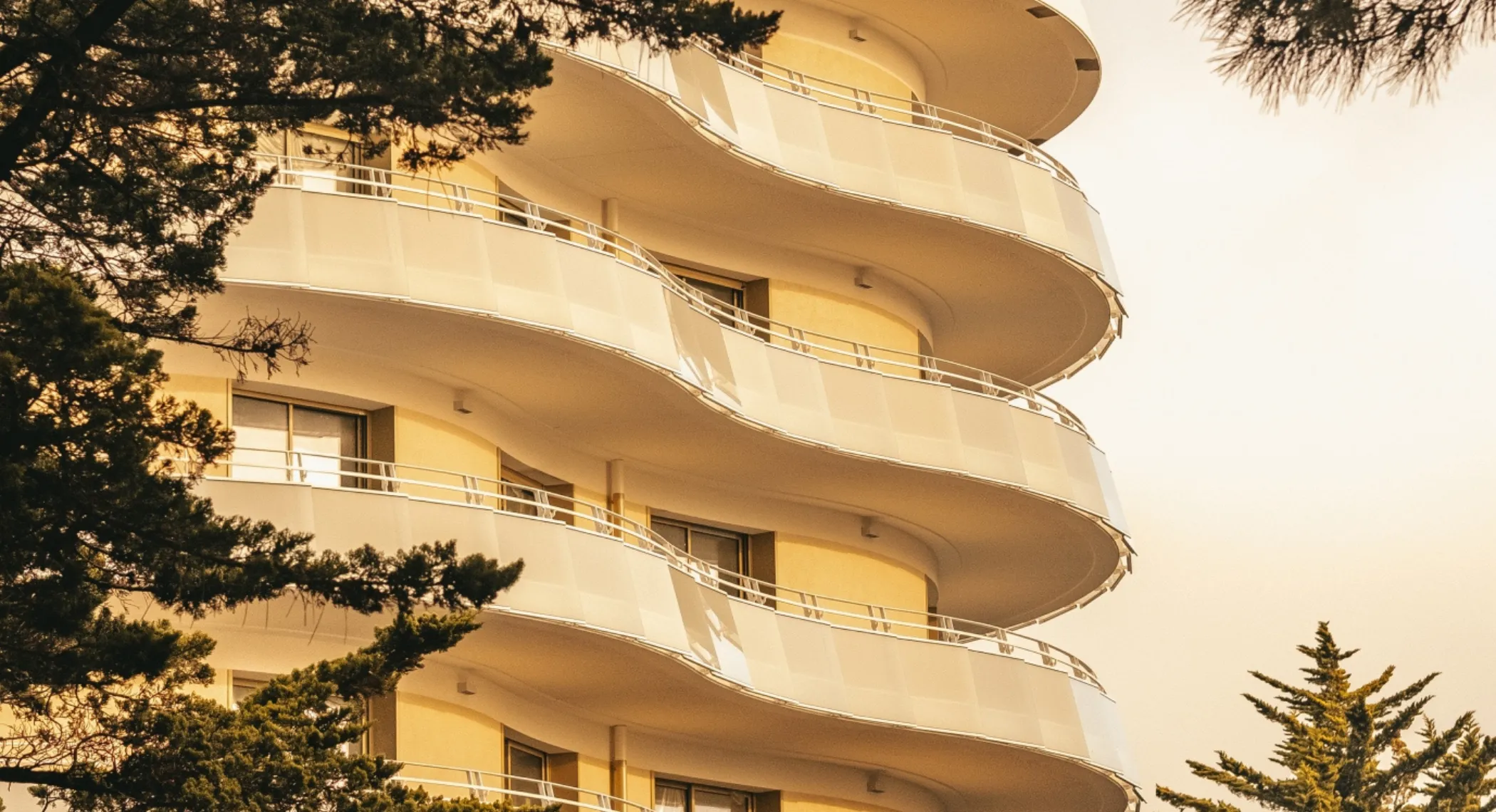 Balcons incurvés blancs d'un immeuble moderne avec des fenêtres et des arbres verts au premier plan.